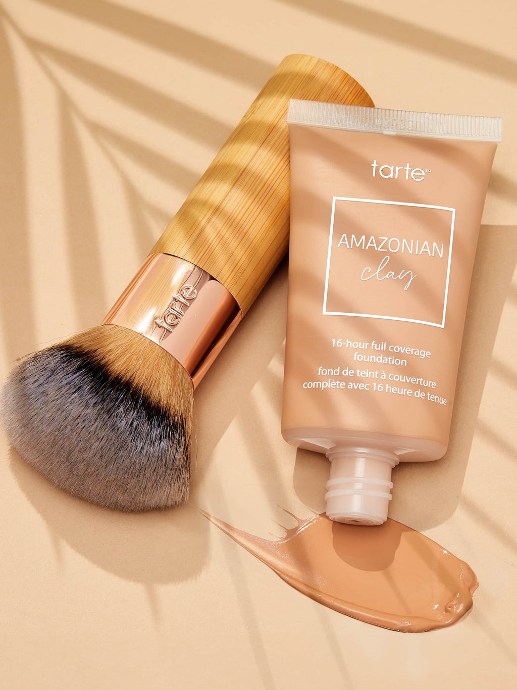 A makeup brush rests beside Tarte Cosmetics Amazonian Clay 16-hour full coverage foundation tube, with some foundation squeezed out, casting soft shadows on a beige surface.